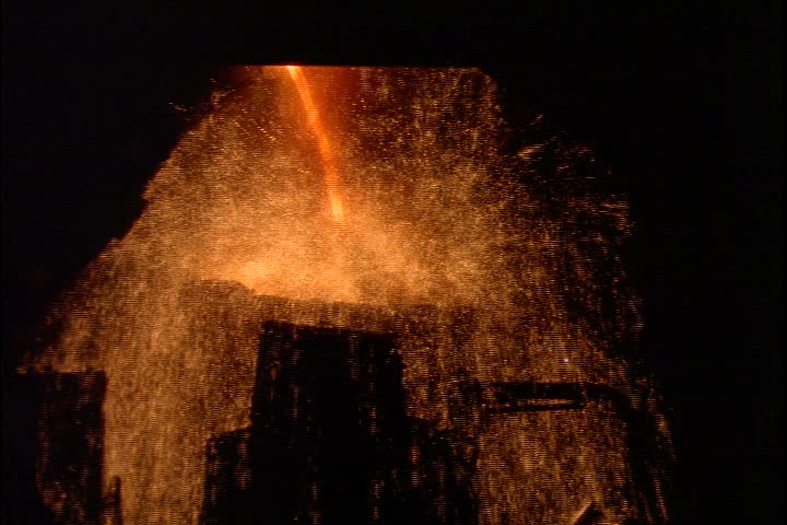 Steel works in Nizhny-Tagil, Russia. Cascade of flames and sparks fills frames. Sparks clear to reveal MS stream of glowing orange molten iron pouring into ladle against black background.