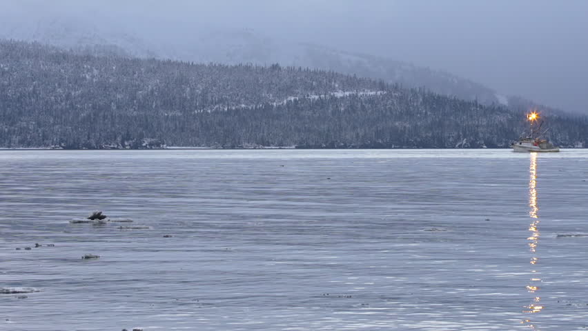 An Alaskan fishing trawler crawls across Kamechak Bay in front of snowy mountains and a forest-covered shore in Homer, Alaska