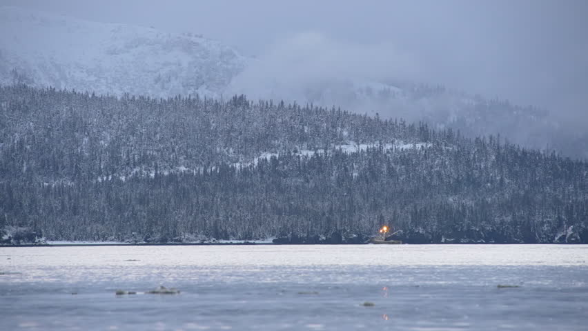 A small fishing trawler passes in front of a dark forest and snowy mountain