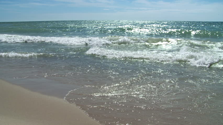 Angled view of waves breaking on a sandy beach on a summer's day.  Looping clip.