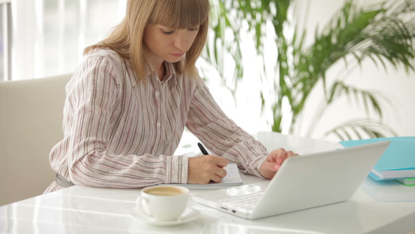 Charming young woman at office table working on laptop writing in notebook and