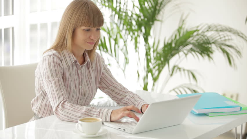 Pretty young businesswoman sitting at table working on laptop and writing in