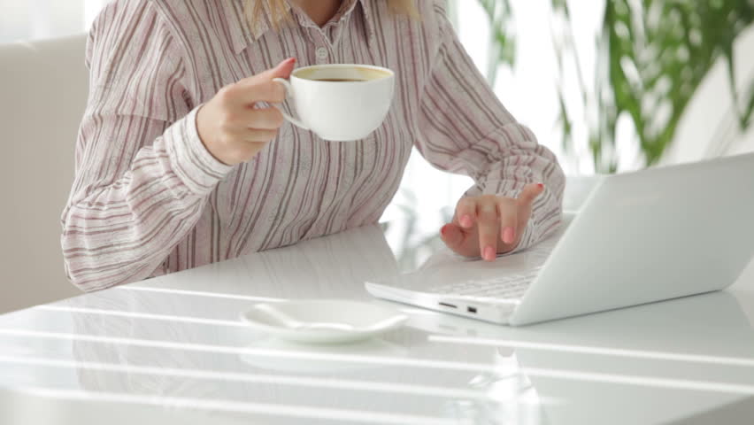 Attractive young woman sitting at table working on laptop drinking coffee and