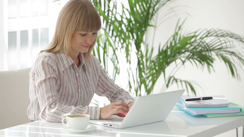 Cute young woman at office working on laptop drinking coffee and smiling
