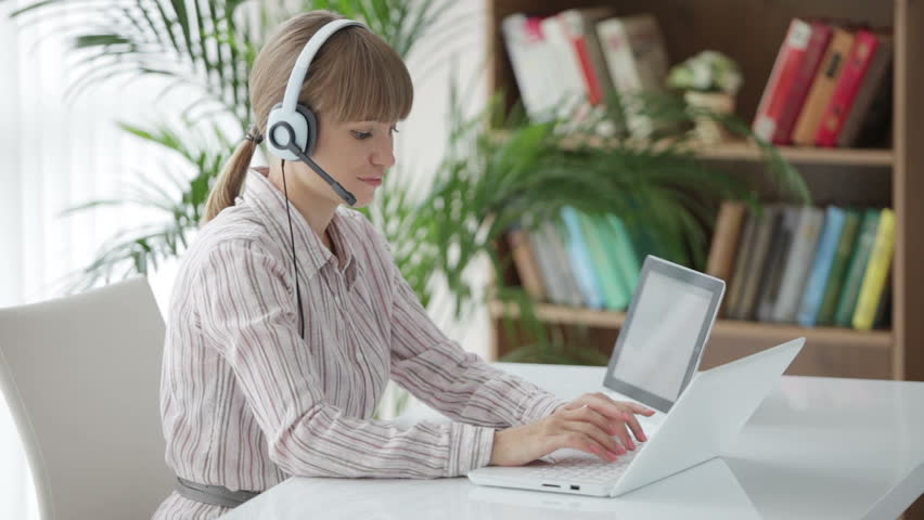 Cheerful young woman wearing headset sitting at table and working on laptop
