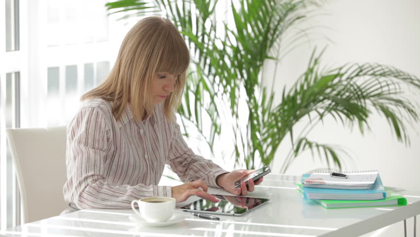 Charming young woman sitting at table using touchpad and mobile phone and