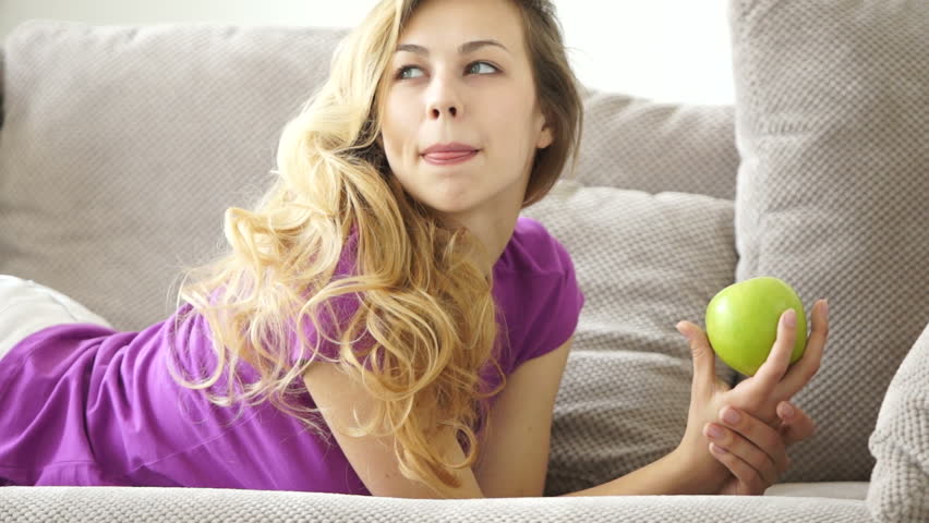 Attractive young woman on sofa holding green apple and smiling at camera
