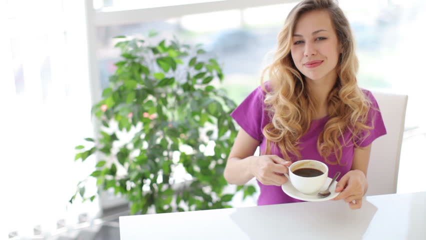 Attractive young woman sitting at table with green plant on background drinking