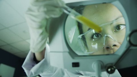 Beautiful Asian laboratory worker is analyzing a liquid sample under the view of a large magnifying glass.