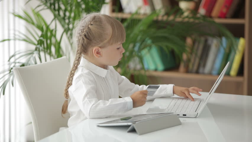 Cute little girl sitting at table holding credit card using laptop and smiling