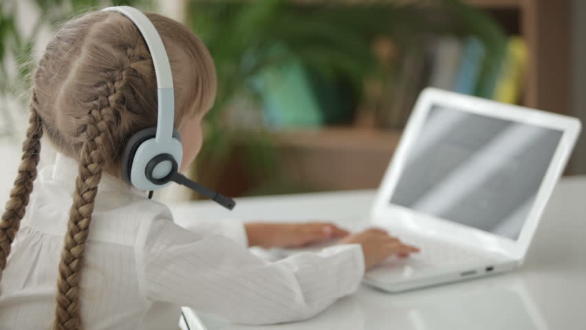 Beautiful little girl in headset with microphone sitting at table using laptop