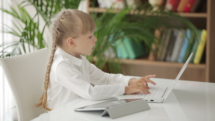 Funny little girl sitting at table with laptop and touchpad and smiling at