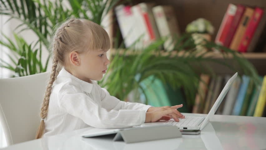 Beautiful little girl sitting at table using laptop and touchpad and smiling at