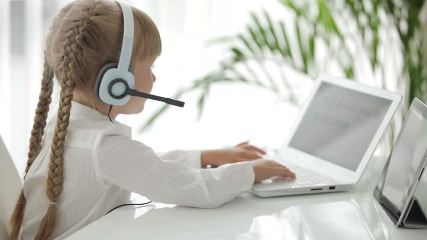 Pretty little girl in headset sitting at table using laptop and touchpad and