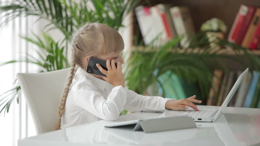 Pretty little girl using laptop talking on mobile phone and looking at camera