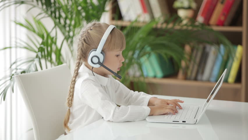 Pretty little girl wearing headset using laptop and smiling at camera
