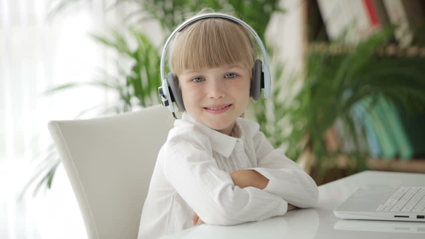 Cute little girl in headset sitting at table with laptop and smiling
