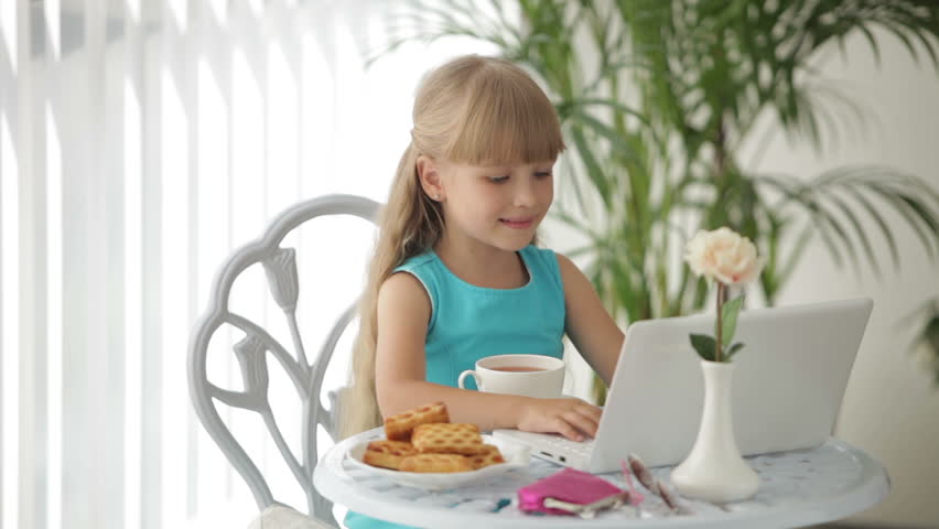 Charming little girl sitting at table with laptop drinking tea and smiling at