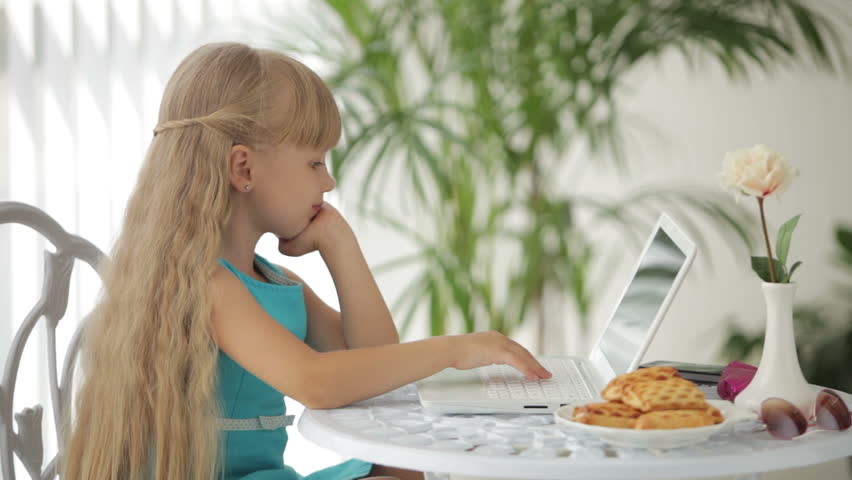 Charming little girl sitting at table with plate of biscuits using laptop and