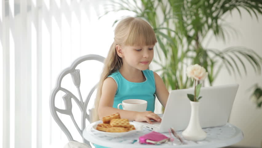 Cute little girl sitting at table with laptop drinking tea and smiling
