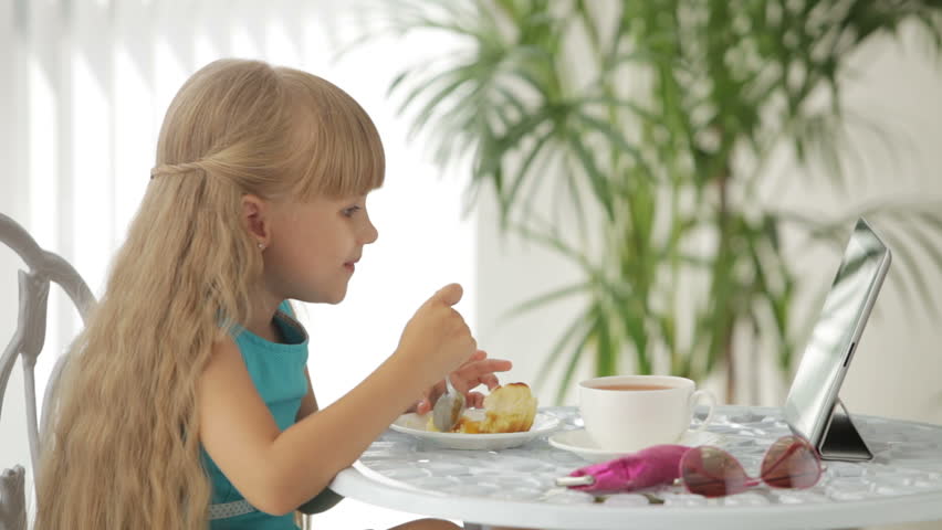 Funny little girl sitting at table eating cake using touchpad and smiling at