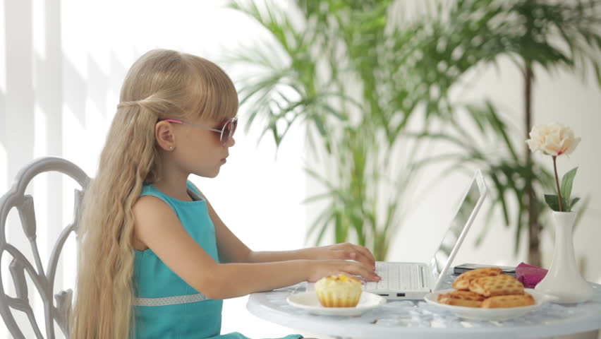 Cute little girl sitting at table using laptop eating cake and smiling at