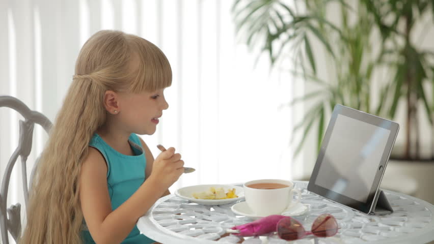 Cute little girl eating cake using touchpad and smiling at camera
