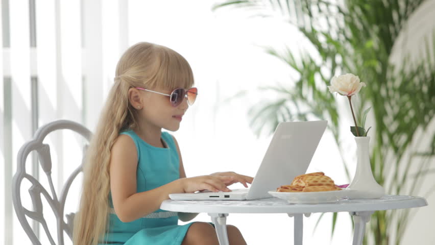 Beautiful little girl wearing sunglasses sitting at table using laptop and