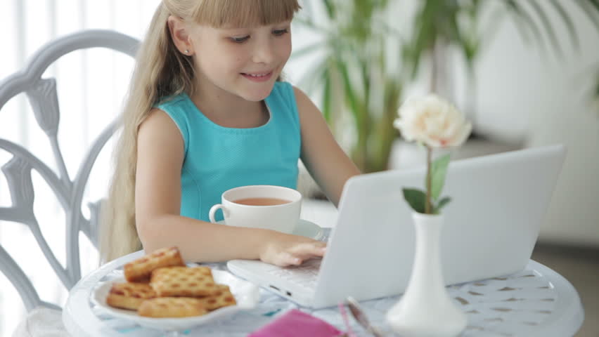 Cute little girl sitting at table with laptop drinking tea showing thumb up and