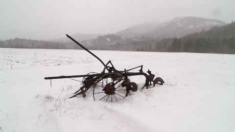 Old antique farm plow stands abandoned in snowy field during a light storm