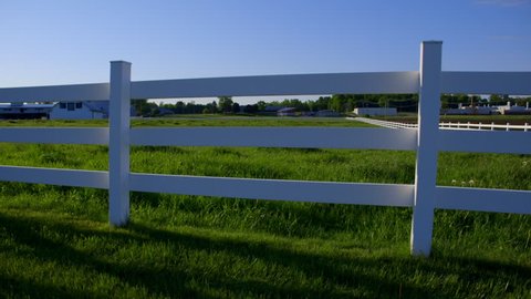 Panning view of white fence with green fields and farmhouse
