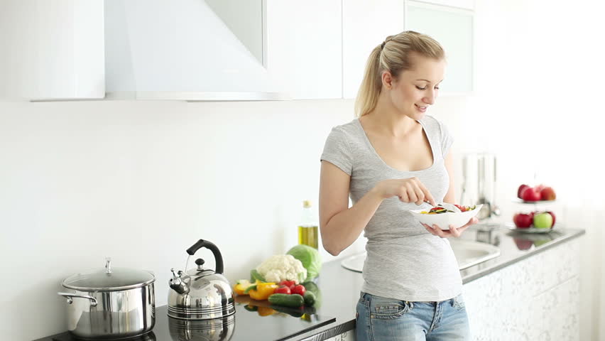 Cheerful young woman in kitchen eating fresh cut vegetable salad from bowl and