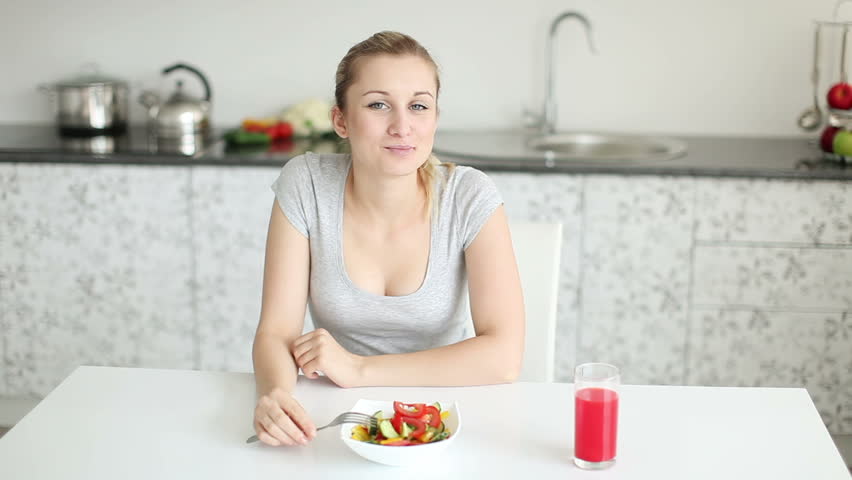 Pretty young woman sitting at kitchen table eating vegetable salad and smiling
