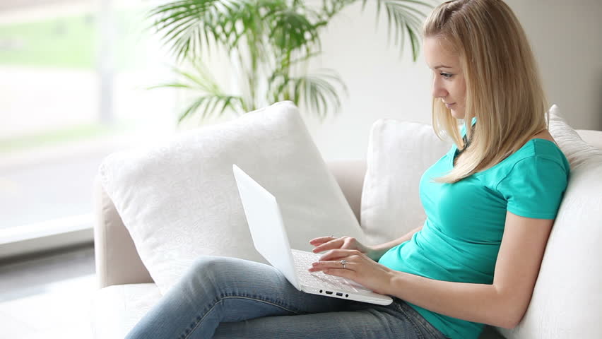 Pretty girl sitting on sofa with laptop on her knees and smiling while typing
