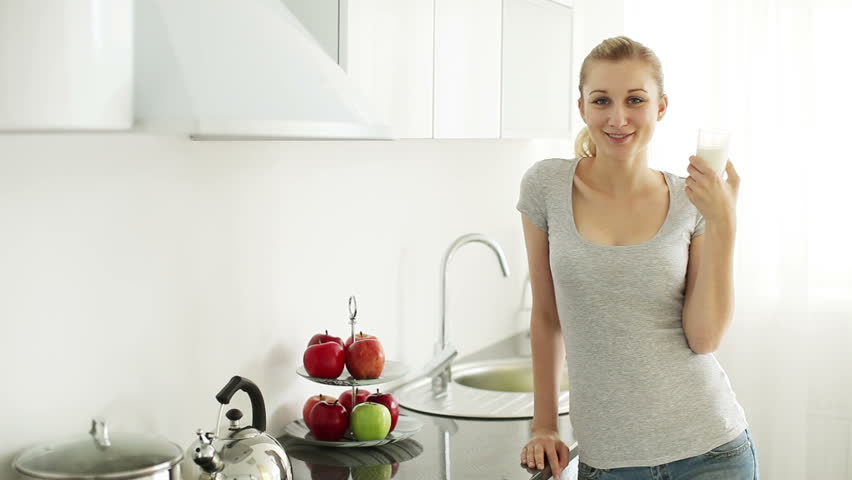 Cheerful young woman standing at kitchen drinking milk from glass and smiling at