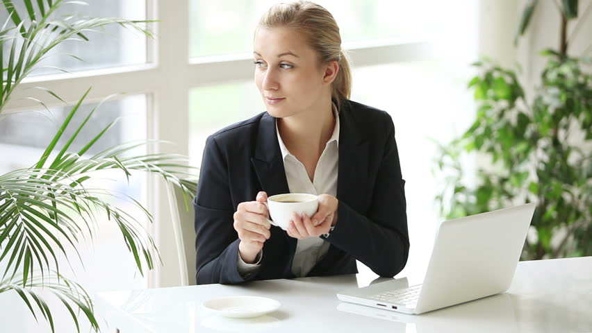 Pretty young woman at office sitting at table holding cup of coffee using laptop