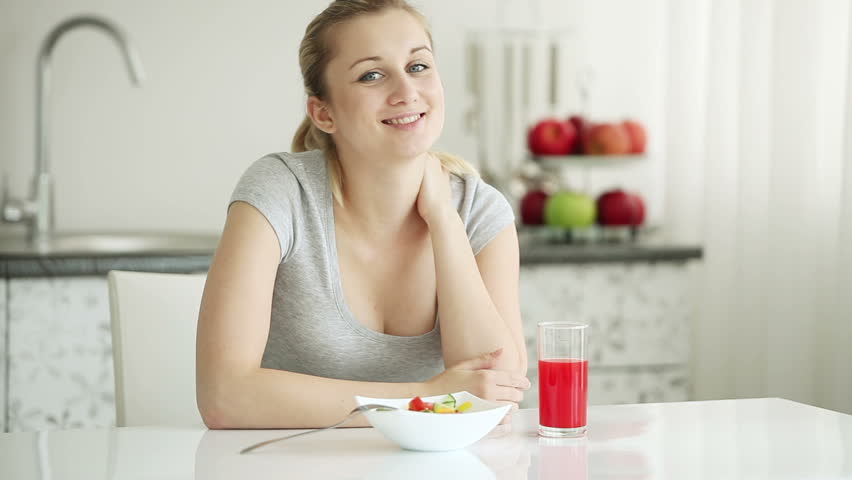 Happy young woman sitting at kitchen table and eating vegetable salad smiling at