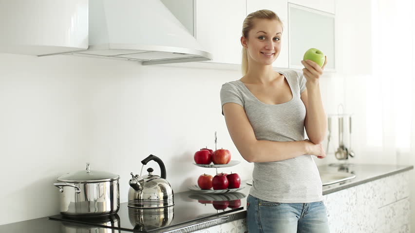 Pretty blond girl standing in kitchen with apple in her hand and smiling at