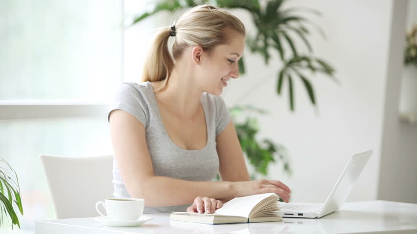 Pretty young woman sitting at table with opened book and laptop and smiling