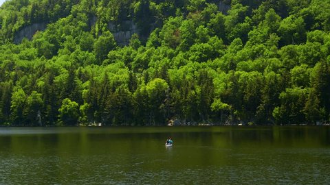 A couple paddles in a canoe, dwarfed by a wooden lakeside cliff
