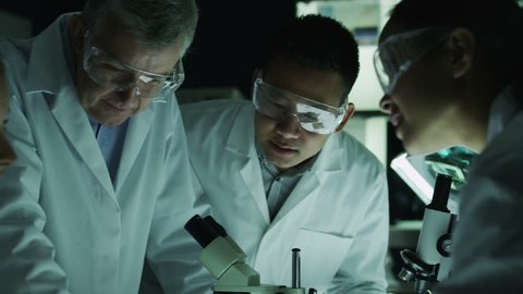 A mature male scientist or medical researcher is working in a dark laboratory, teaching a young team of students or trainees. In slow motion.