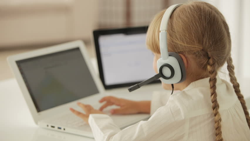 Pretty little girl sitting at desk with headset using laptop looking at camera