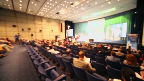 People sit on rows of chairs in large hall during conference, (defocused)