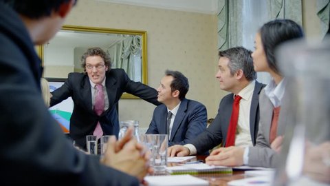 Happy and enthusiastic business team of mixed ages and ethnicity are seated around a conference table for a business meeting. They reach across the table to shakes hands with one another. Slow motion.