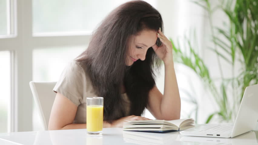 Beautiful young woman sitting at table with glass of juice reading book and