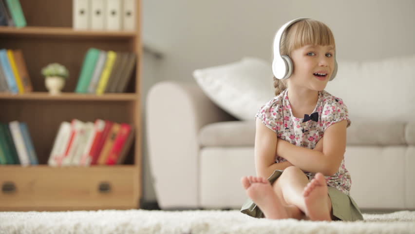 Pretty little girl in headphones sitting on carpet in living room singing to