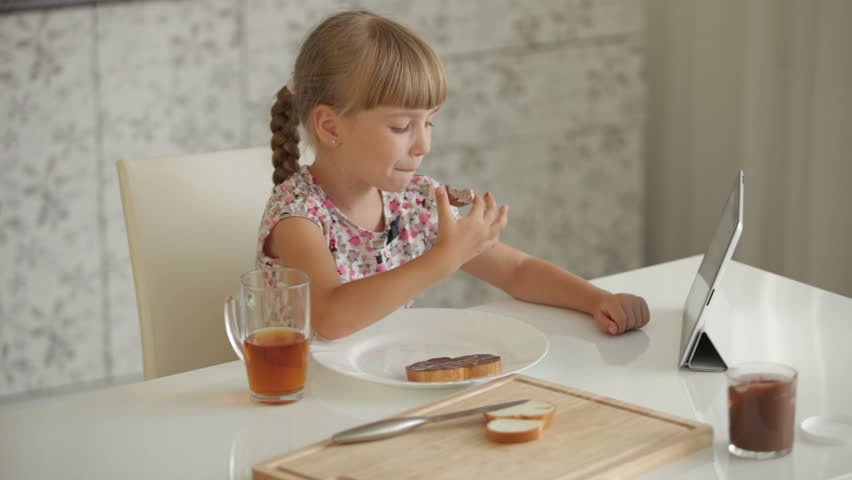 Pretty little girl sitting at kitchen table eating chocolate sandwich and using
