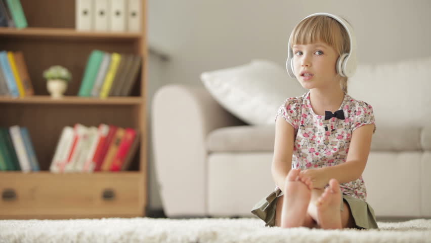 Funny little girl in headphones sitting on carpet in living room singing, moving