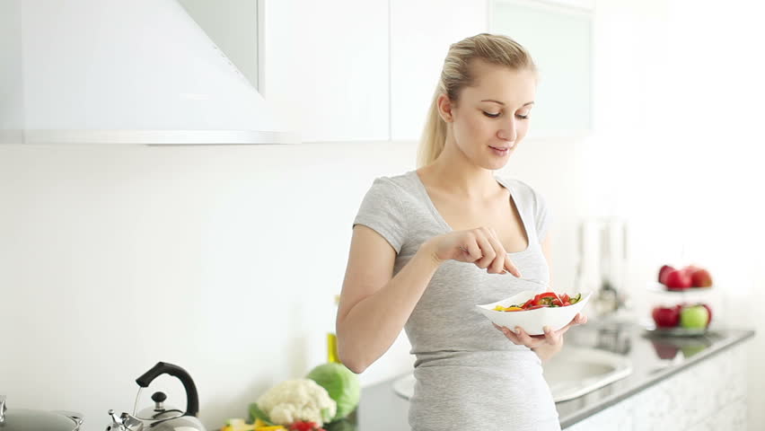 Attractive young woman in kitchen eating vegetable salad and smiling happily