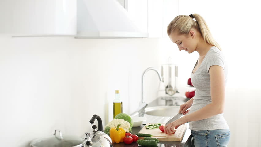 Attractive young woman standing at kitchen table cutting tomatoes looking at